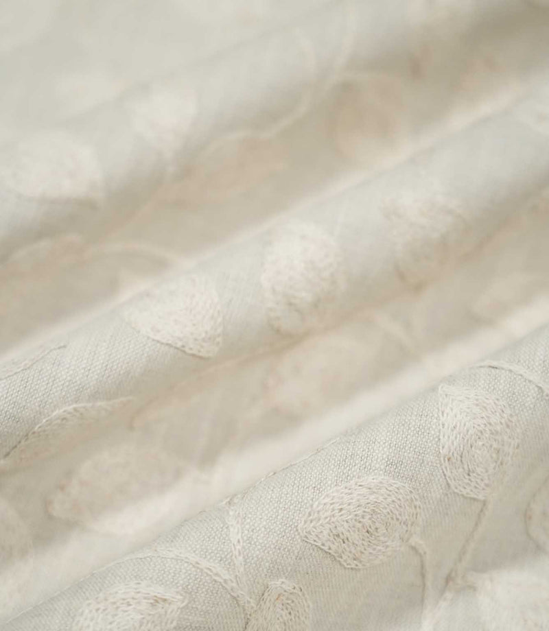 Close-up of Ellerton Cloud embroidered fabric arranged in gentle flowing folds, showing tonal ivory botanical leaves and trailing stems across a pale cloud-grey woven ground, with selective focus highlighting the raised embroidery and natural texture while the surrounding fabric softly fades out of focus.