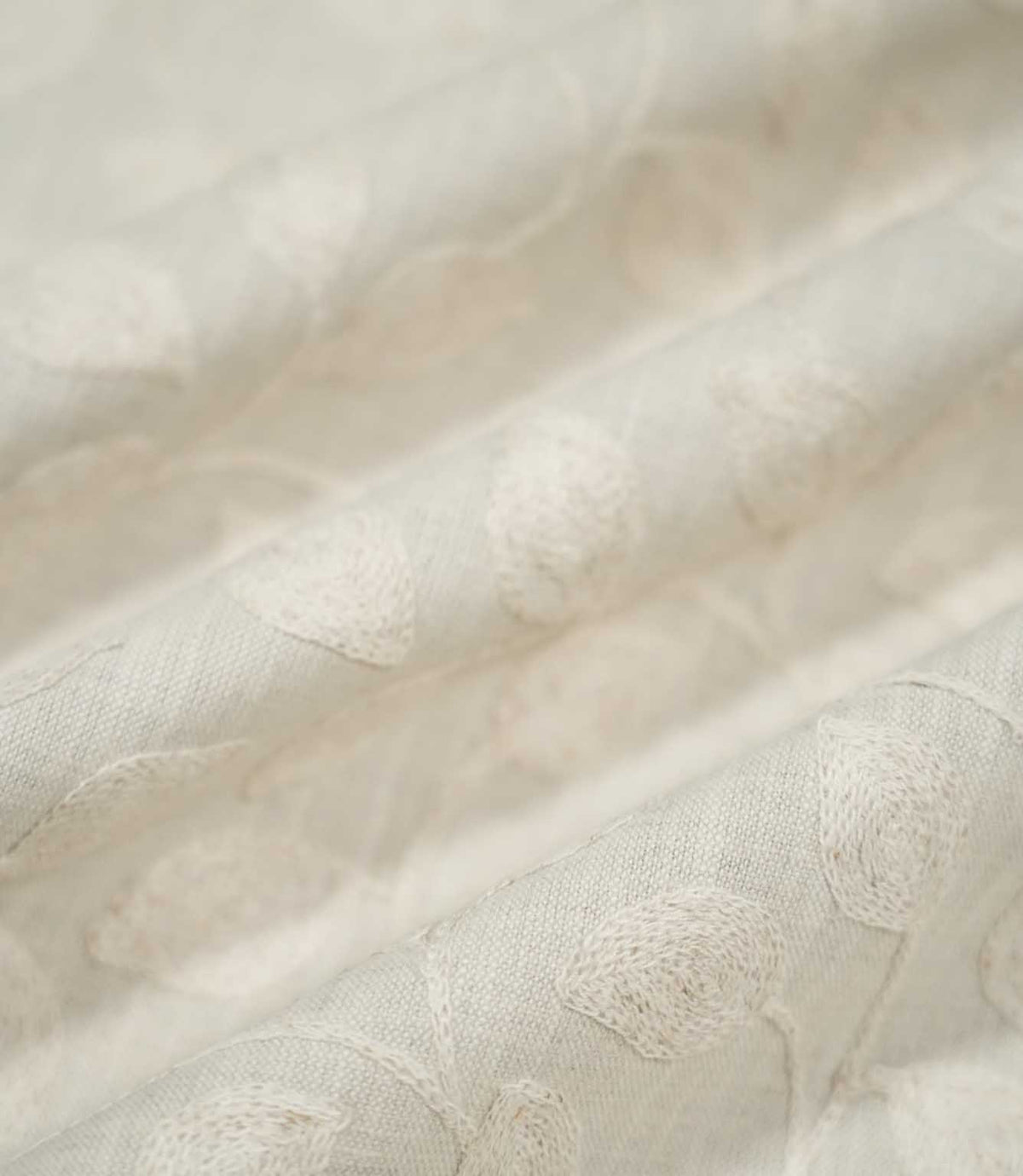 Close-up of Ellerton Cloud embroidered fabric arranged in gentle flowing folds, showing tonal ivory botanical leaves and trailing stems across a pale cloud-grey woven ground, with selective focus highlighting the raised embroidery and natural texture while the surrounding fabric softly fades out of focus.
