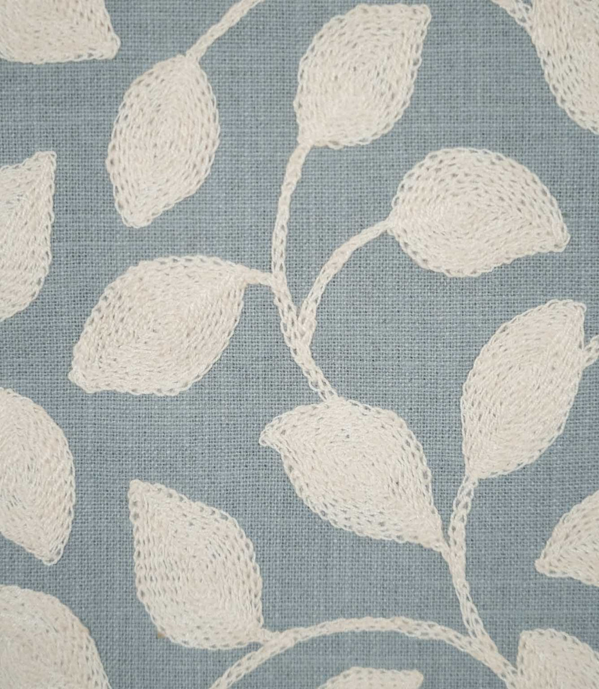 Detailed close-up of Ellerton Cornflower fabric showing raised cream leaf embroidery and delicate stitched stems against a textured cornflower blue woven ground, highlighting the individual threads and tactile finish of the contemporary foliage design.
