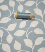 Ellerton Cornflower embroidered foliage fabric shown with a coordinating blue thread spool, featuring textured cream leaves and trailing stems across a soft cornflower blue background, with the close view highlighting the fabric weave and embroidery.