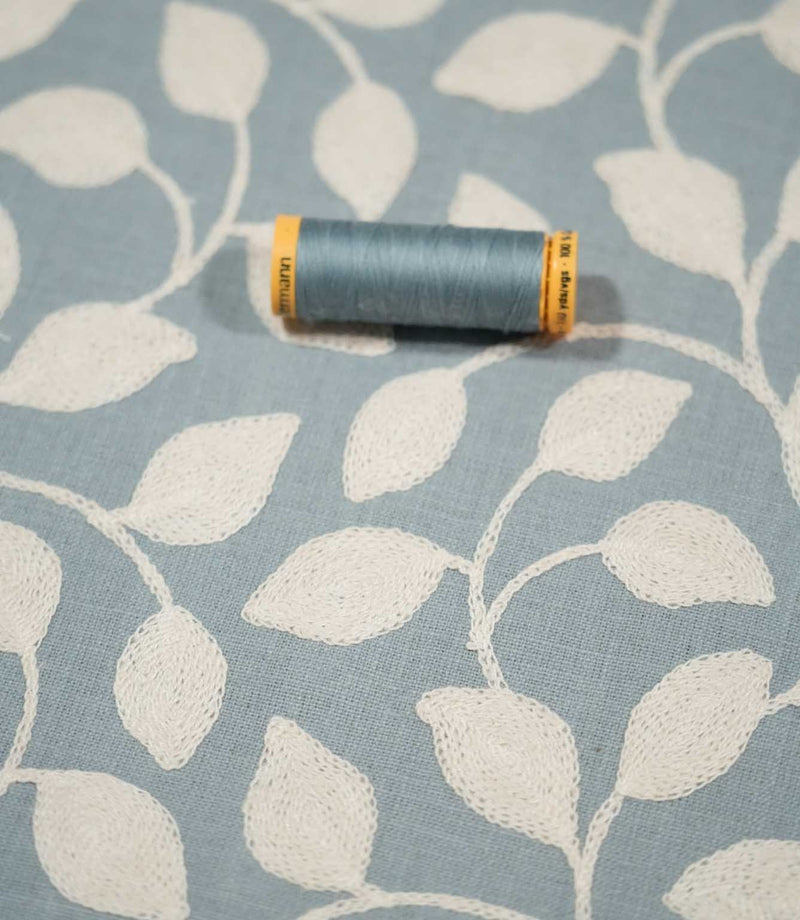 Ellerton Cornflower embroidered foliage fabric shown with a coordinating blue thread spool, featuring textured cream leaves and trailing stems across a soft cornflower blue background, with the close view highlighting the fabric weave and embroidery.