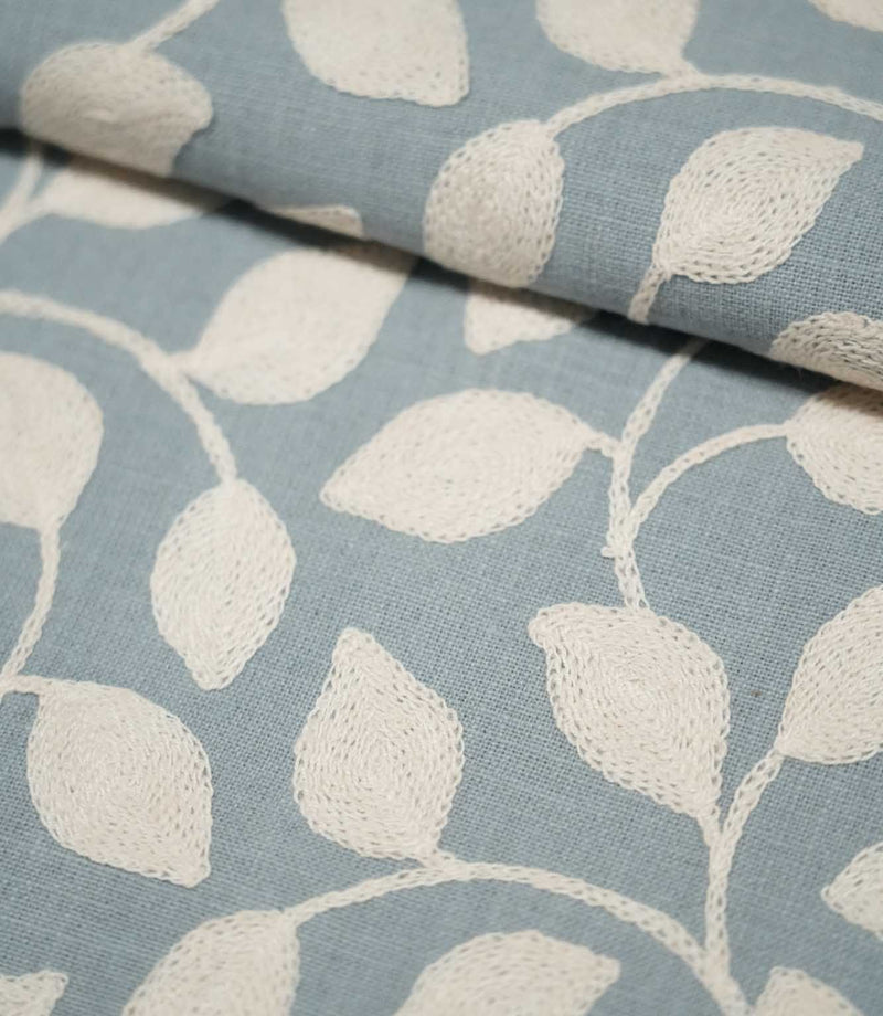 Close-up of folded Ellerton Cornflower fabric showing the cream embroidered foliage pattern across a cornflower blue woven background, with the raised leaf stitching and curved stems clearly visible across the smooth fold.