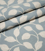 Ellerton Cornflower embroidered fabric arranged in neat overlapping folds, displaying the repeating cream leaf and trailing stem design against a cornflower blue background while highlighting the fabric's woven texture, embroidered surface and structure.
