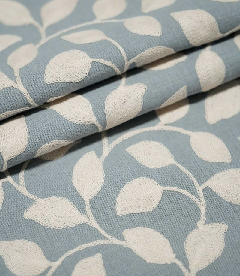 Ellerton Cornflower embroidered fabric arranged in neat overlapping folds, displaying the repeating cream leaf and trailing stem design against a cornflower blue background while highlighting the fabric's woven texture, embroidered surface and structure.