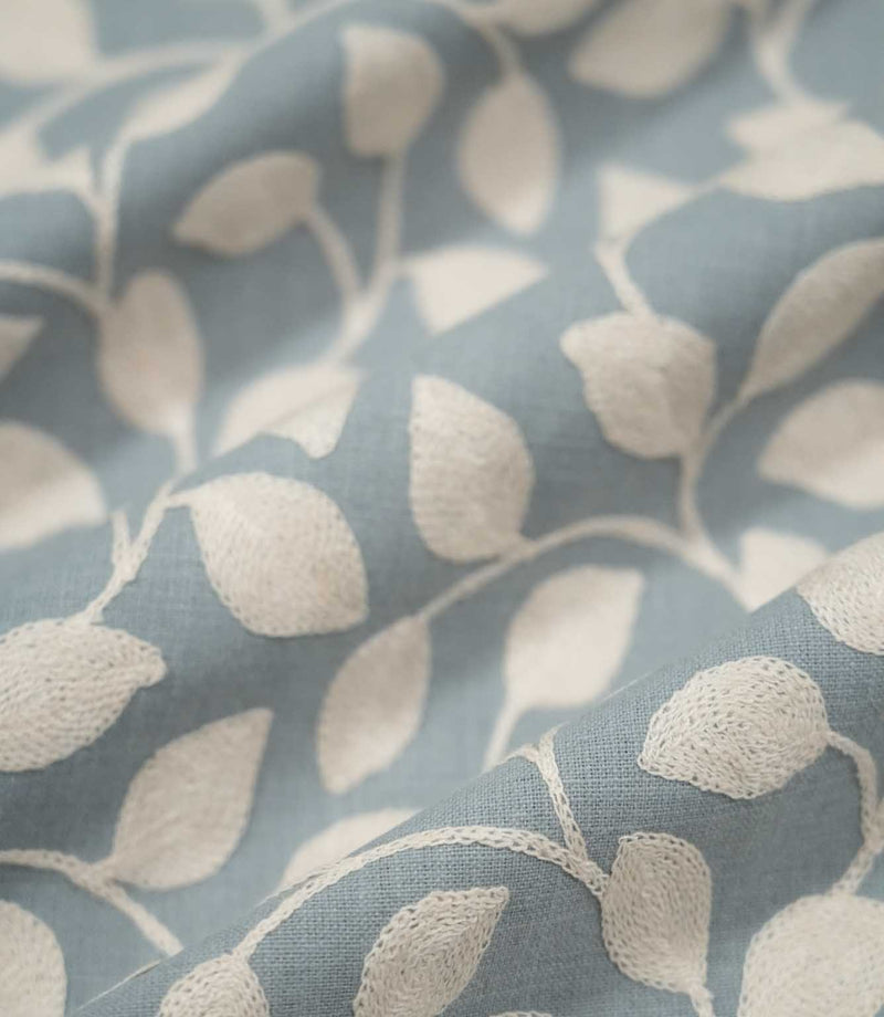 Ellerton Cornflower embroidered fabric arranged in gentle flowing folds, with cream foliage embroidery sweeping across the cornflower blue woven ground, showing the botanical pattern, textured stitching and soft drape suitable for curtains, blinds, cushions and decorative upholstery.