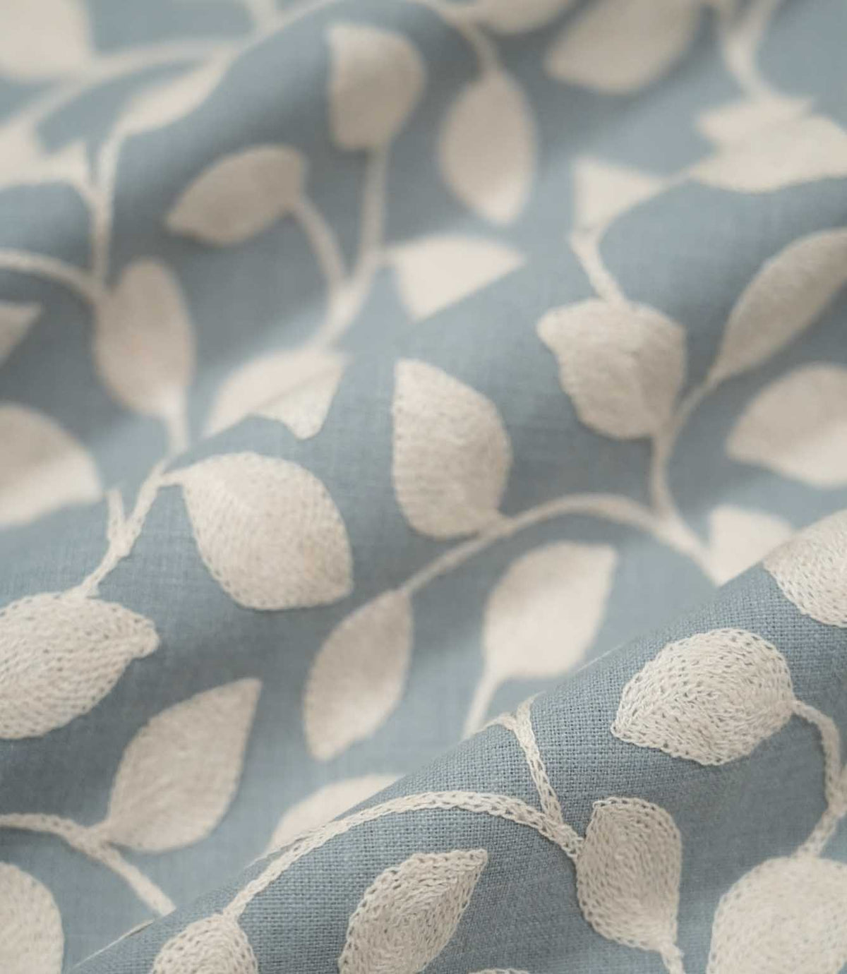 Ellerton Cornflower embroidered fabric arranged in gentle flowing folds, with cream foliage embroidery sweeping across the cornflower blue woven ground, showing the botanical pattern, textured stitching and soft drape suitable for curtains, blinds, cushions and decorative upholstery.