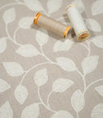 Ellerton embroidered foliage fabric with cream and natural beige sewing thread spools resting on the surface, showing the neutral woven ground and softly textured cream leaf embroidery.