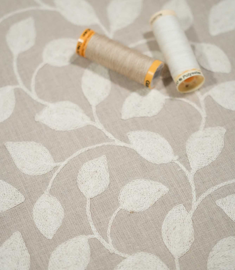 Ellerton embroidered foliage fabric with cream and natural beige sewing thread spools resting on the surface, showing the neutral woven ground and softly textured cream leaf embroidery.