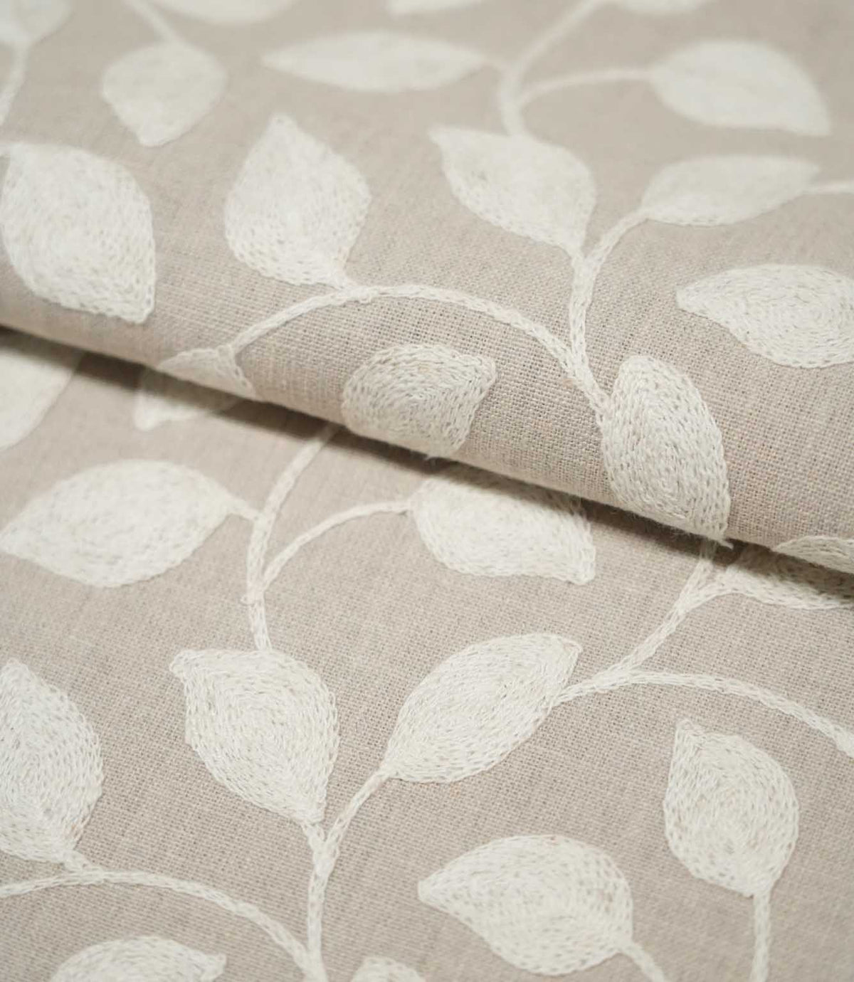 Close-up of folded Ellerton fabric showing cream embroidered leaves and flowing stems across a natural beige background, with the fold highlighting the fabric's woven texture and embroidered surface.