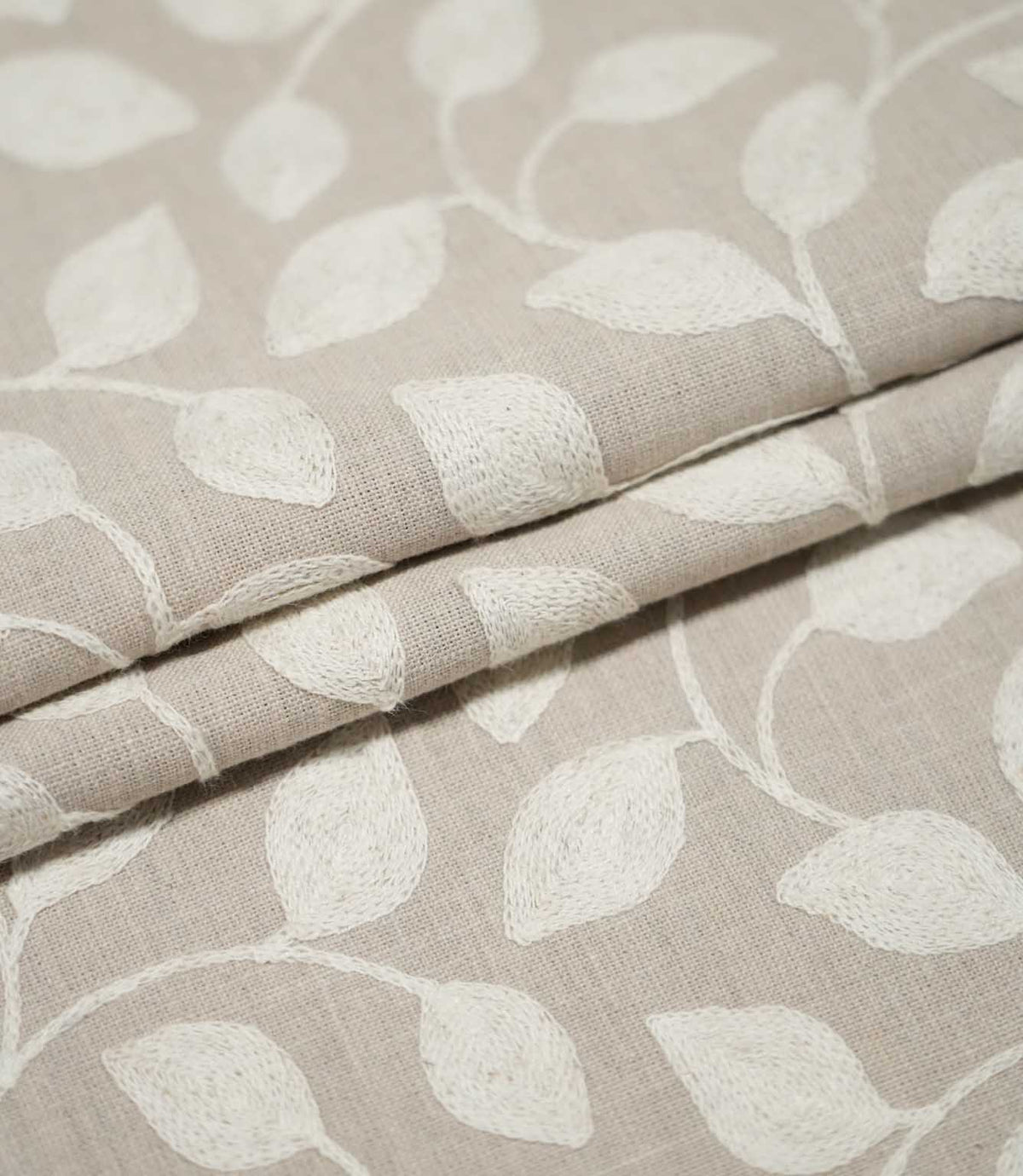 Ellerton fabric arranged in neat layered folds, displaying the repeating cream embroidered foliage design and natural beige woven ground while showing the fabric's thickness and texture.