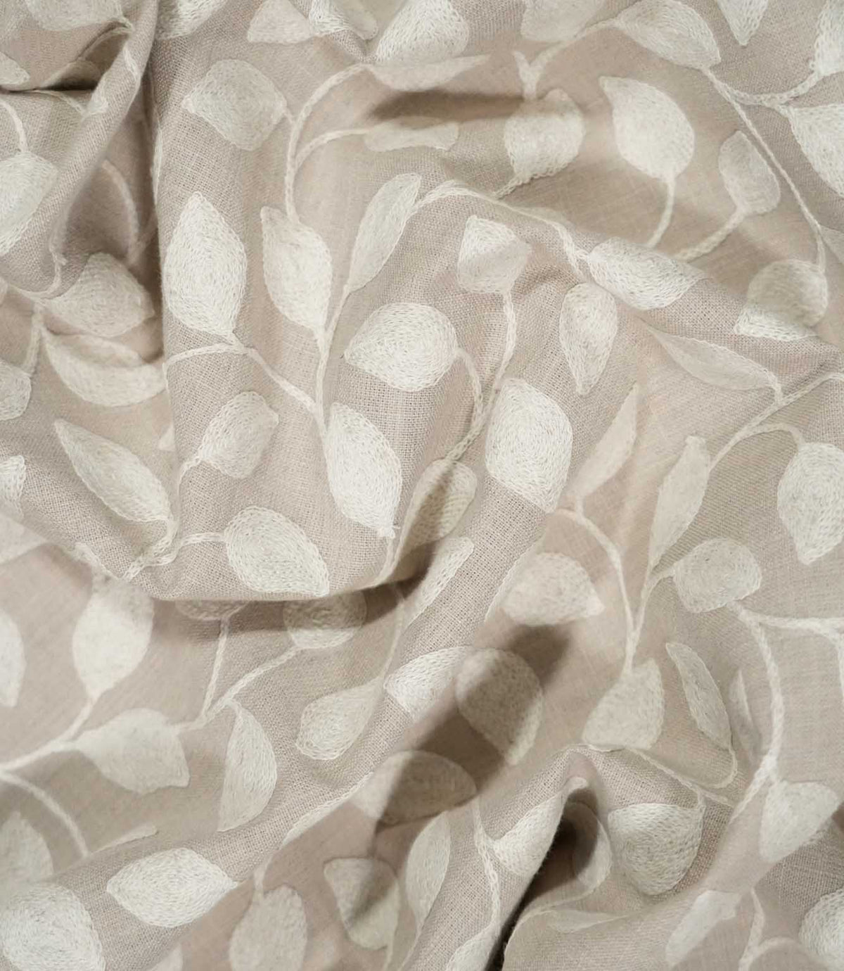 Ellerton embroidered fabric loosely draped into natural folds, showing how the cream trailing leaf pattern moves across the natural beige ground and demonstrating the fabric's soft handle and drape.