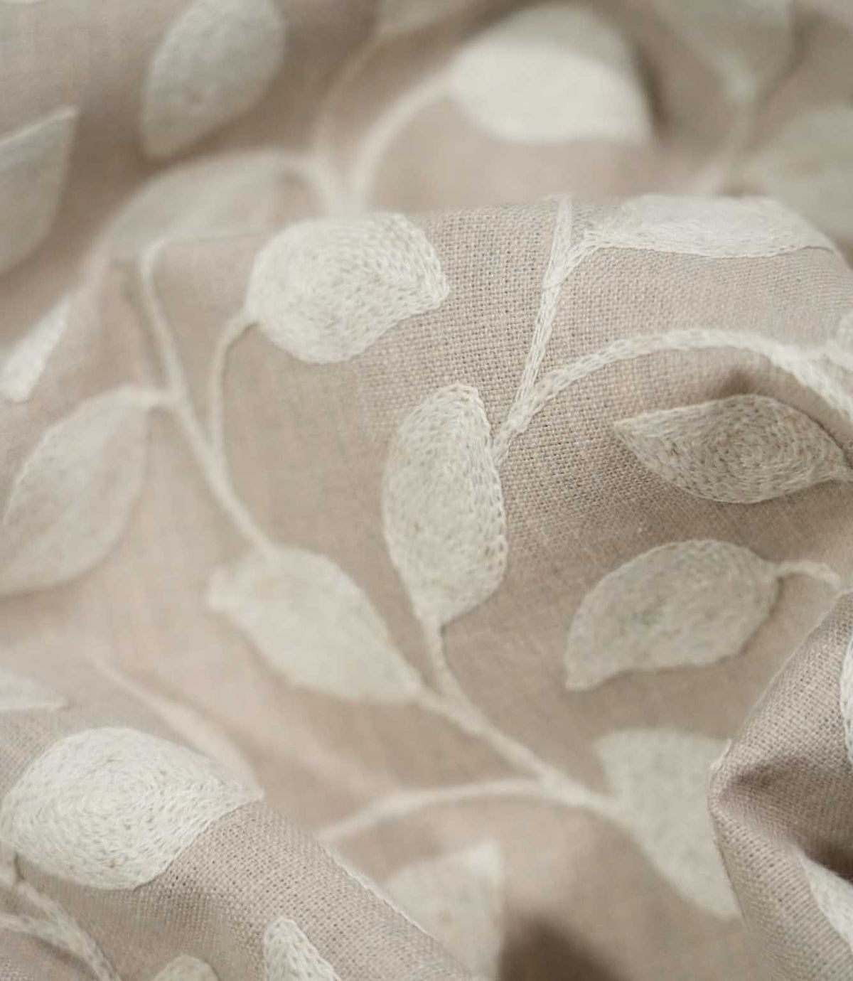 Detailed close-up of Ellerton fabric gathered into soft folds, highlighting the raised cream leaf embroidery, winding stems and fine texture of the natural beige woven background.
