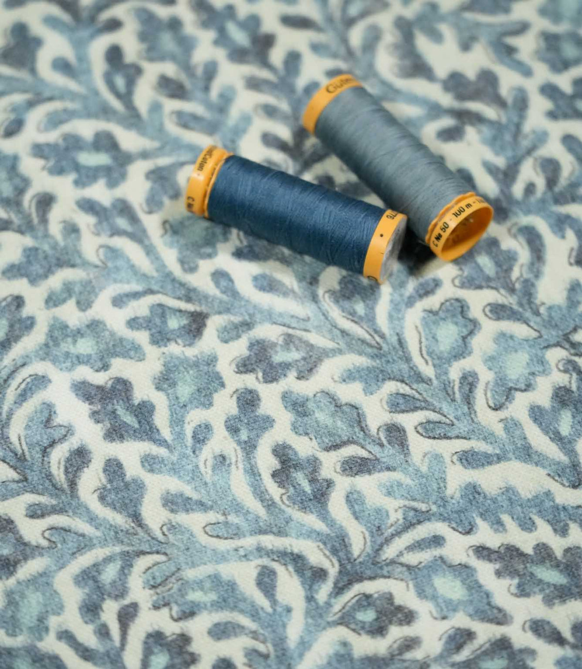 Elmsley blue floral fabric with two coordinating blue thread spools resting on the surface, showing the fabric's detailed print, texture and tonal blue palette.