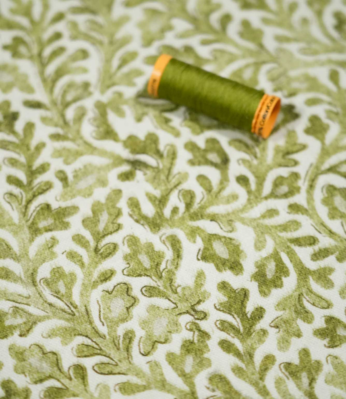 Elmsley Green floral fabric displayed with a coordinating olive green thread spool, showing the detailed foliage pattern, softly textured print and natural green tones against an off-white ground.