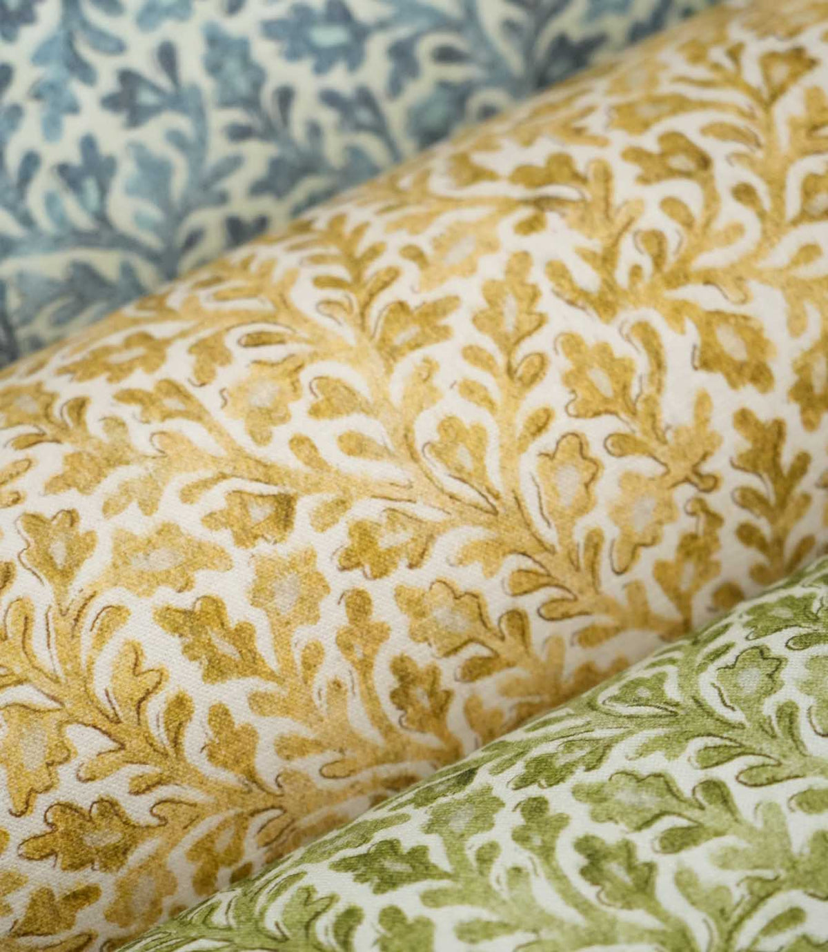 Close-up of the Elmsley fabric collection layered together, showing the Ochre colourway alongside coordinating Green and Blue designs, all featuring the same small-scale botanical foliage and floral print on an off-white ground.
