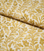 Close-up of folded Elmsley Ochre fabric showing the botanical pattern flowing naturally over the fold, with stylised leaves and flowers in warm ochre, mustard and golden tones on an off-white background.
