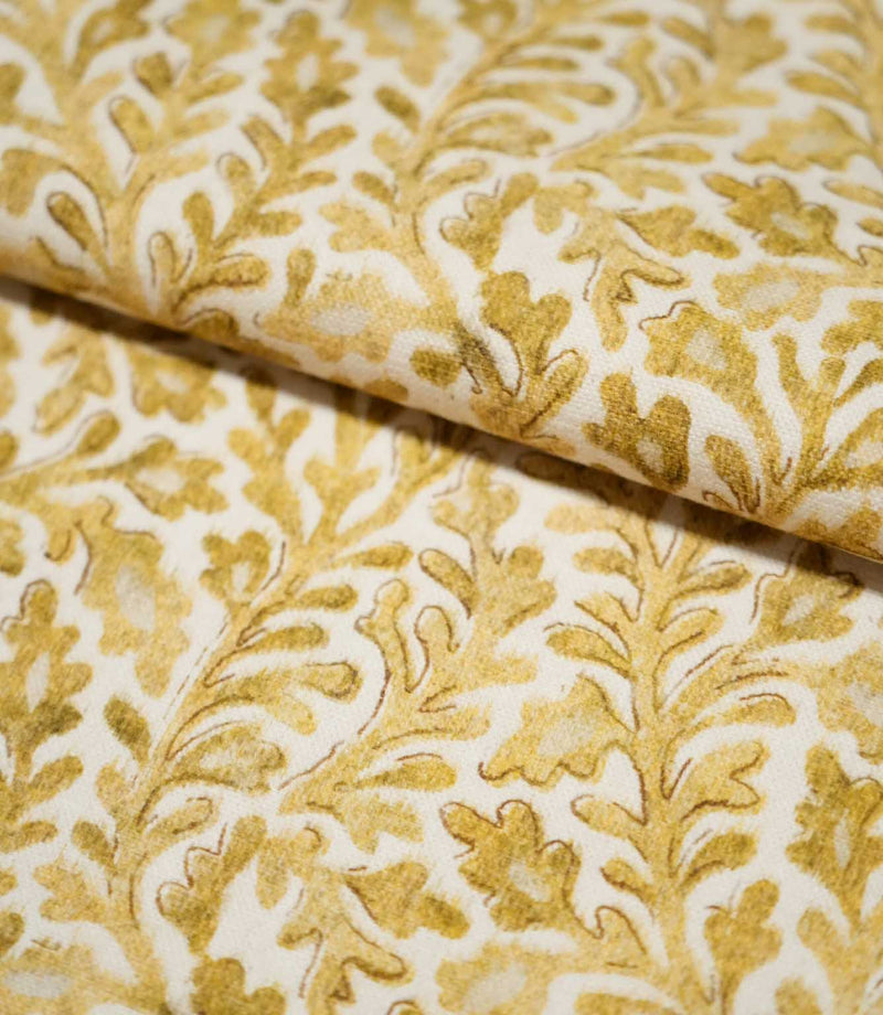 Close-up of folded Elmsley Ochre fabric showing the botanical pattern flowing naturally over the fold, with stylised leaves and flowers in warm ochre, mustard and golden tones on an off-white background.