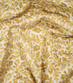 Elmsley Ochre fabric loosely draped into soft, flowing folds, showing the movement and scale of its intricate botanical design with warm golden ochre leaves, stems and stylised flowers across an off-white ground.