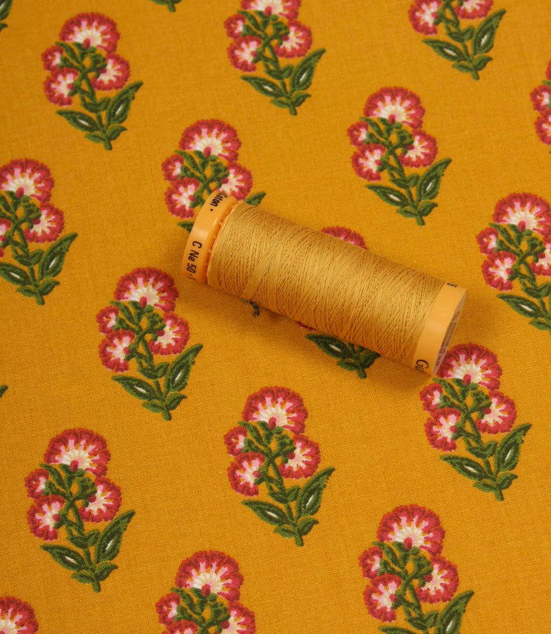 Floral print fabric in mustard yellow shown with a matching thread spool, featuring pink flower motifs and green leaves.
