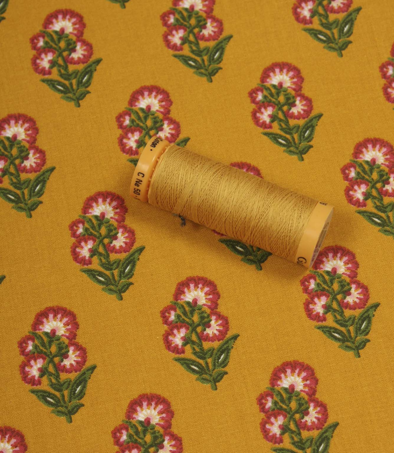 Floral print fabric in mustard yellow shown with a matching thread spool, featuring pink flower motifs and green leaves.