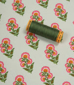 Floral cotton-style fabric shown with coordinating green thread spool, featuring repeating pink blossom motifs on an ivory background.