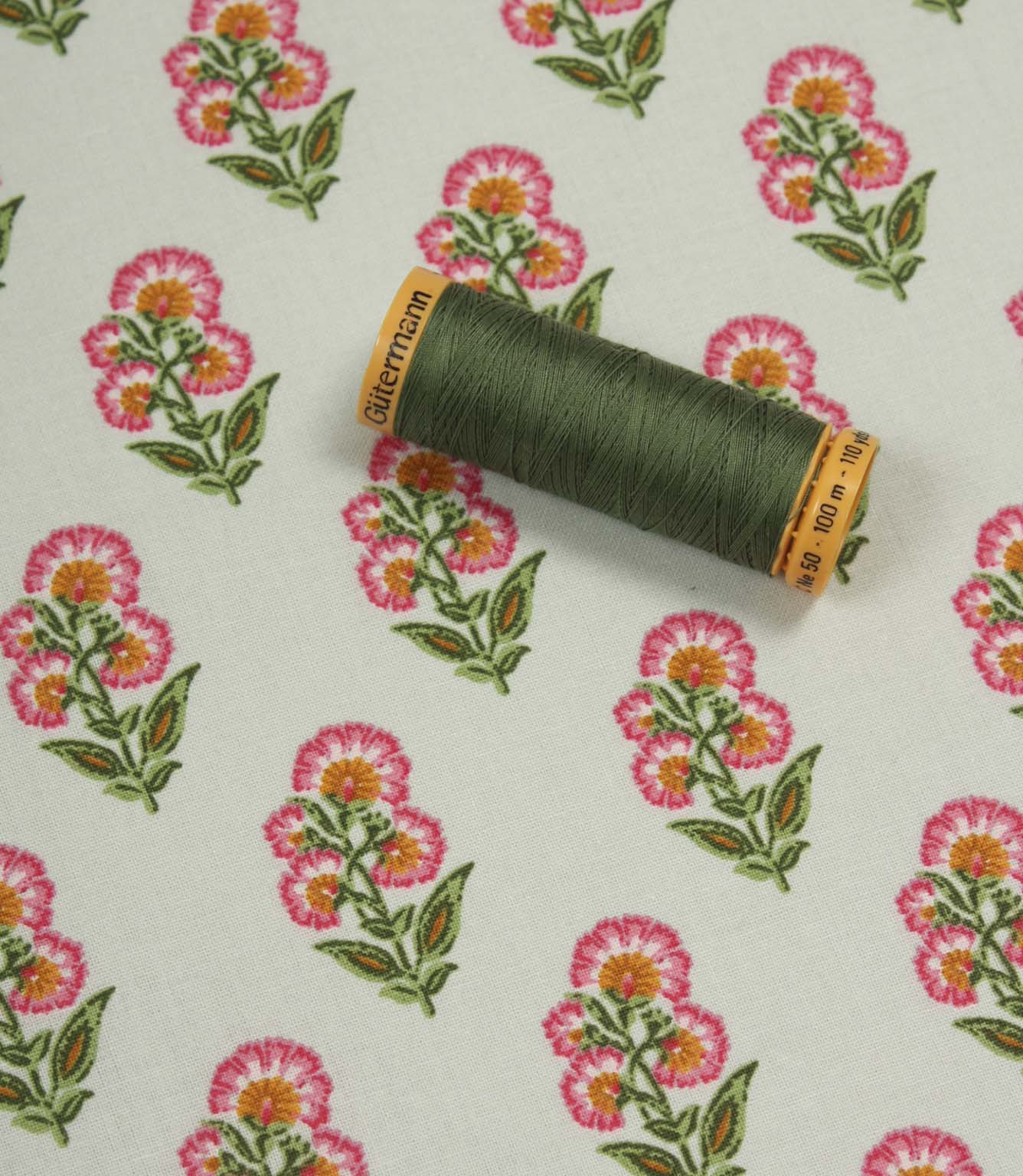 Floral cotton-style fabric shown with coordinating green thread spool, featuring repeating pink blossom motifs on an ivory background.