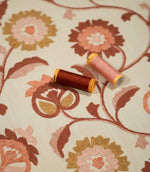 Detailed view of Foxmere Burnt Sienna fabric with two coordinating thread spools resting on the embroidered floral design, highlighting the raised burnt sienna and pink stitching, ochre accents and natural woven base cloth.