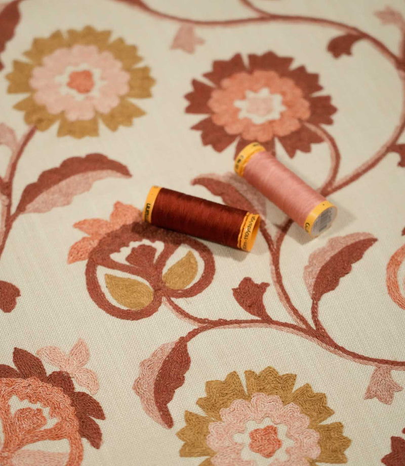 Detailed view of Foxmere Burnt Sienna fabric with two coordinating thread spools resting on the embroidered floral design, highlighting the raised burnt sienna and pink stitching, ochre accents and natural woven base cloth.