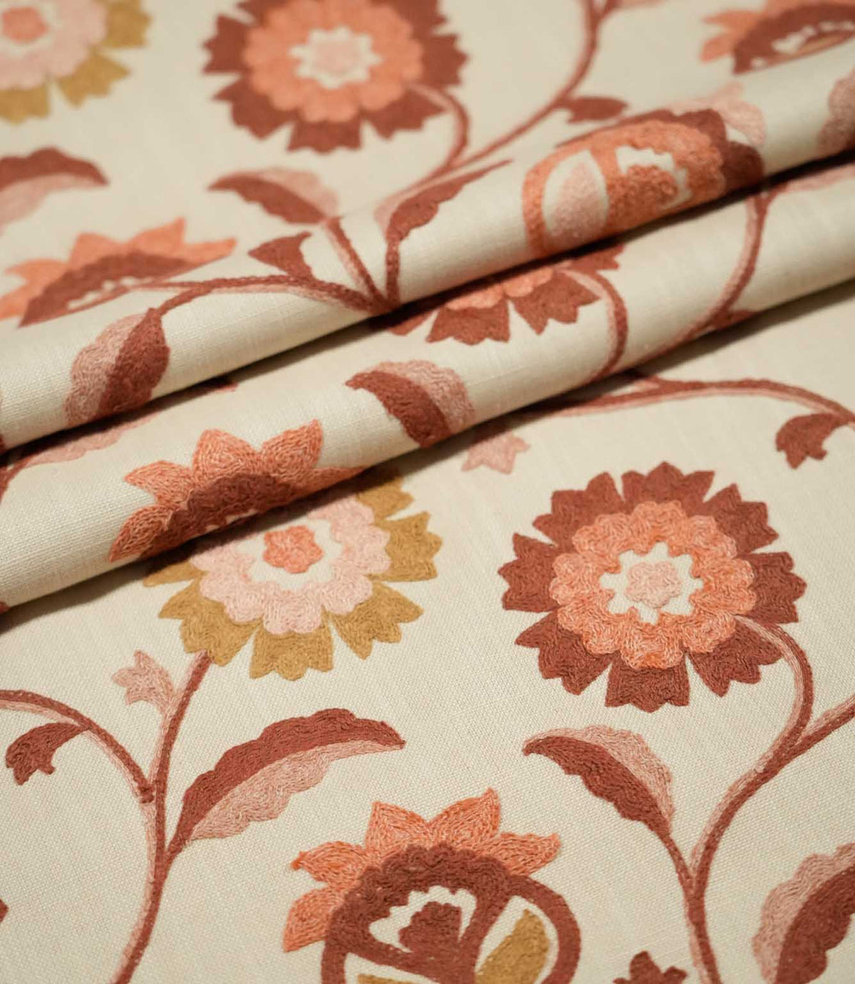 Foxmere Burnt Sienna fabric arranged in soft folds, displaying the flowing embroidered vine pattern with large stylised flowers, leaves and buds in earthy burnt sienna, terracotta, blush pink and ochre tones.