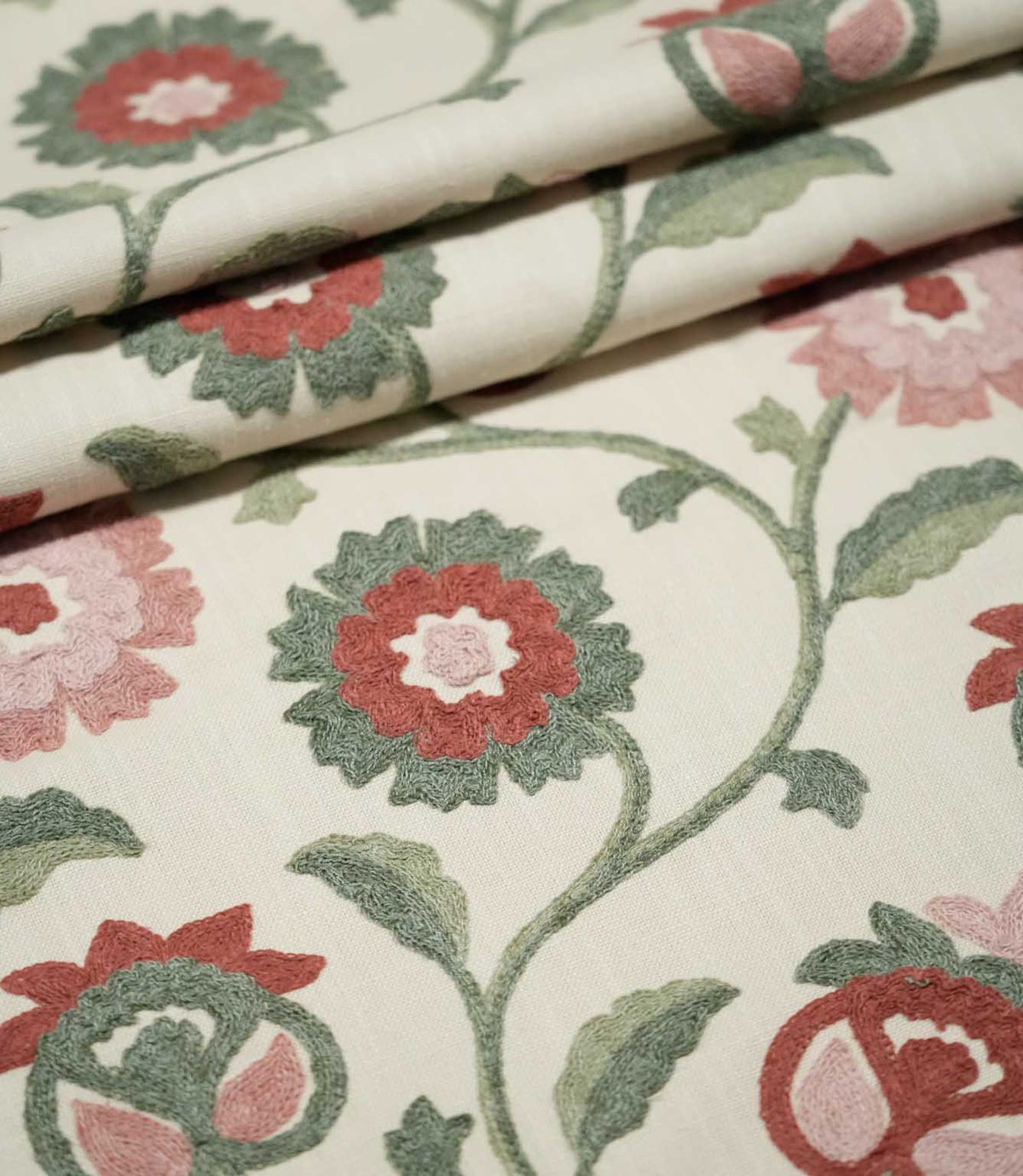Foxmere Chintz fabric arranged in gentle folds, displaying its repeating embroidered botanical design of curving green vines, two-tone leaves and stylised flowers and buds in soft pink, muted red and sage green.