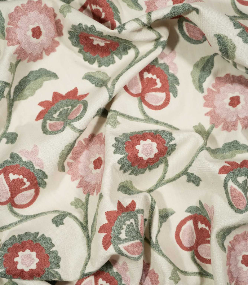 Foxmere Chintz embroidered fabric loosely draped into soft folds, showing how the decorative floral pattern falls across the cloth, with pink and muted red flowers connected by flowing sage green stems and leaves on a natural cream background.