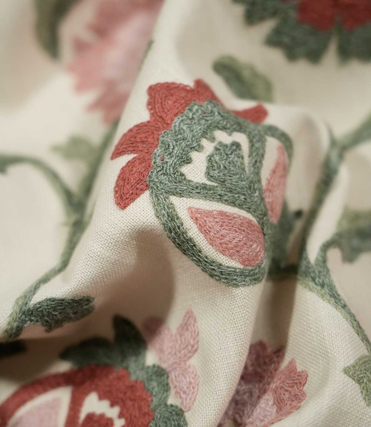 Macro close-up of Foxmere Chintz floral fabric draped into a fold, highlighting the raised embroidery of a stylised flower bud with sage green outlines, dusty pink petals and muted red details against the finely woven cream base cloth.