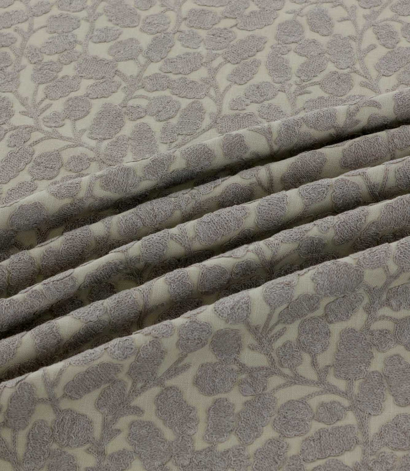 Close-up of Gardiner Gull fabric arranged in soft folds, showing the raised grey embroidered foliage pattern on a natural cream background and its textured finish.