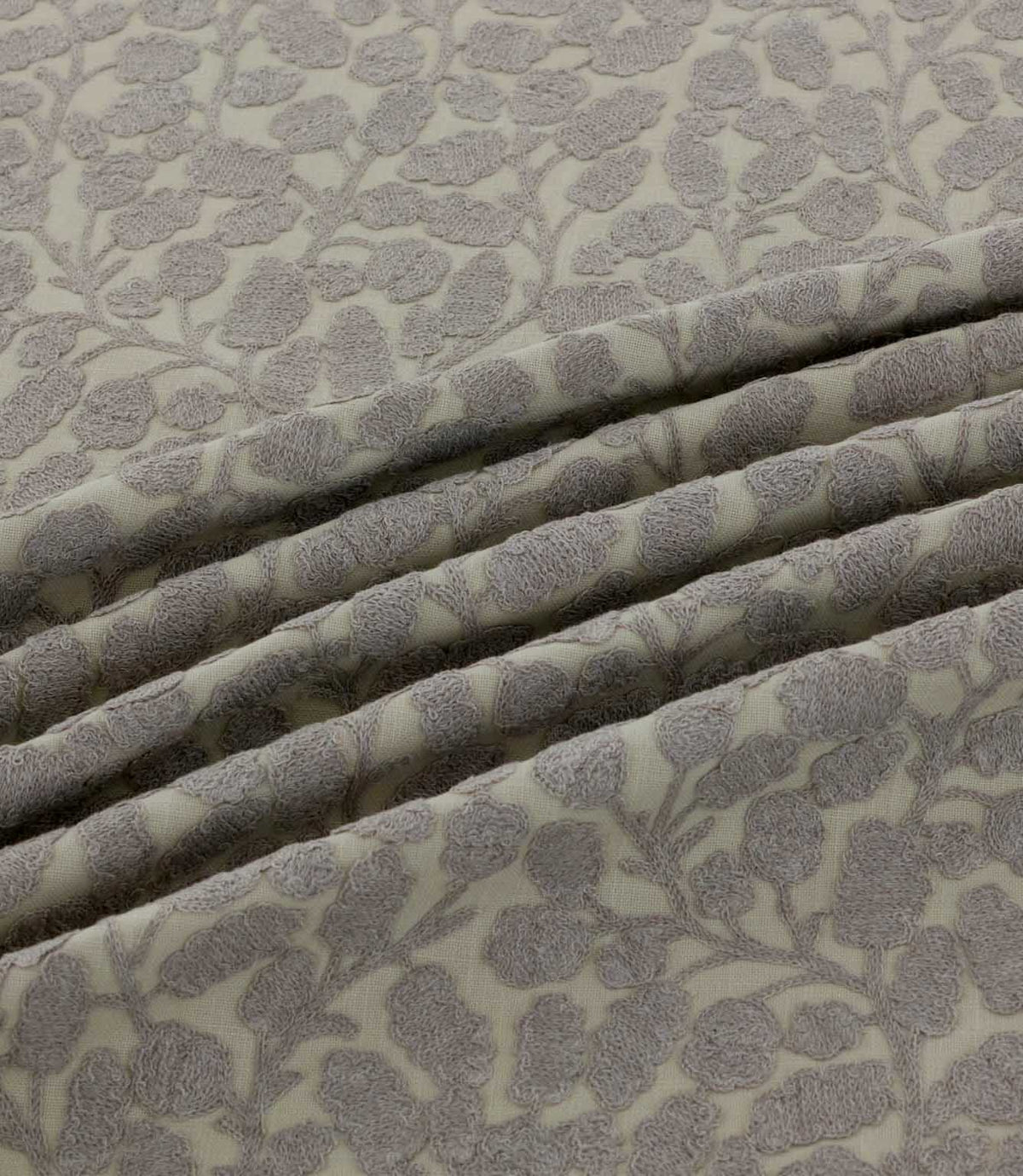 Close-up of Gardiner Gull fabric arranged in soft folds, showing the raised grey embroidered foliage pattern on a natural cream background and its textured finish.