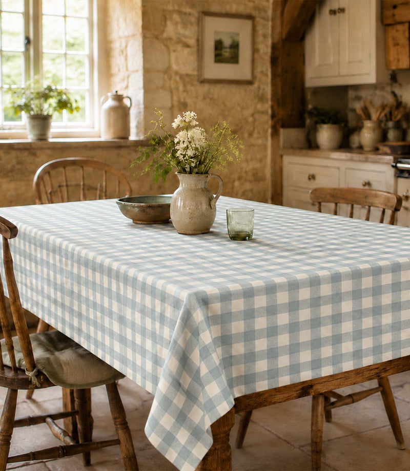 A light blue and white gingham checkered tablecloth draped over a wooden dining table in a rustic kitchen setting, styled with a ceramic vase of white flowers, a small glass, and a metal bowl.