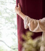 A person gently holds a deep beetroot red Heath washed velvet curtain beside a bright window, showing its soft, subtly textured surface.