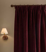Close view of deep beetroot red Heath washed velvet pencil pleat curtains hanging from a dark metal pole beside a softly lit wall lamp.