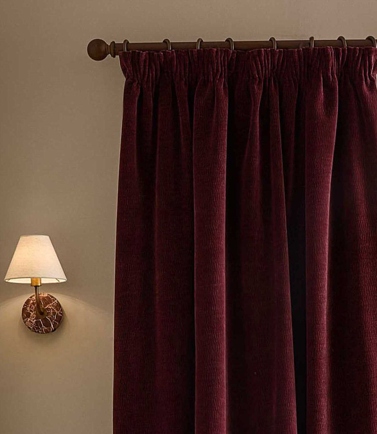 Close view of deep beetroot red Heath washed velvet pencil pleat curtains hanging from a dark metal pole beside a softly lit wall lamp.