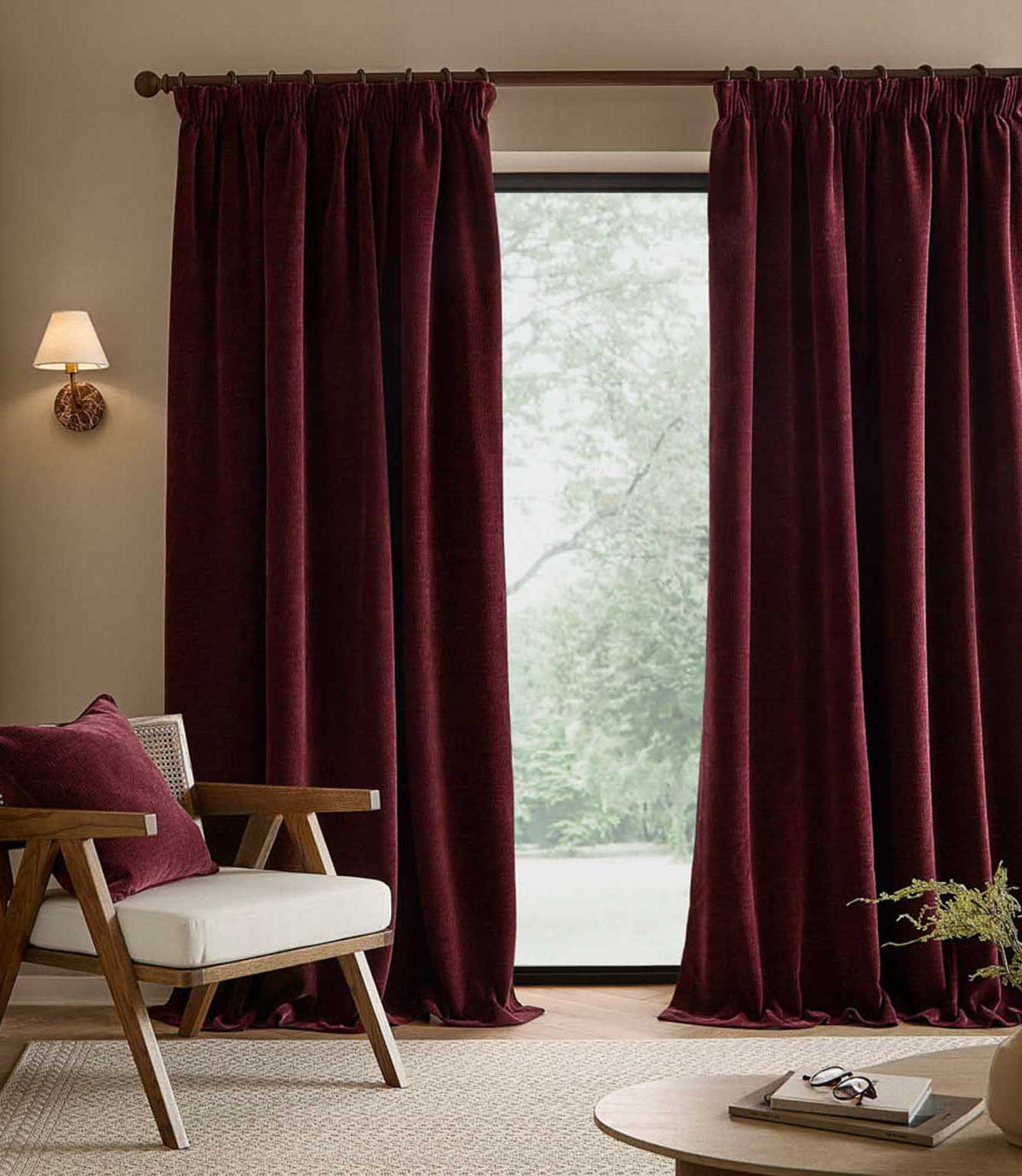 Full-length deep beetroot red Heath washed velvet curtains frame a large window in a neutral living room with a wooden armchair and matching cushion.
