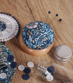 William Morris-inspired pincushion with a navy blue, turquoise, cream and gold floral fabric top and wooden base, holding three navy-headed sewing pins on a craft table.