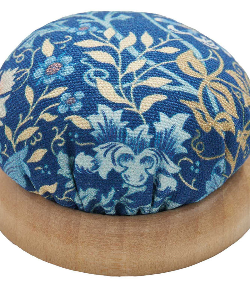 Close-up of a William Morris-inspired pincushion with a rounded wooden base and padded navy blue fabric top decorated with intricate blue, cream and gold flowers and foliage.