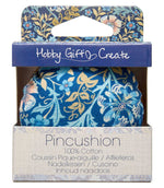 William Morris-inspired floral pincushion in retail packaging, featuring a navy blue, turquoise, cream and gold botanical pattern and natural wooden base.