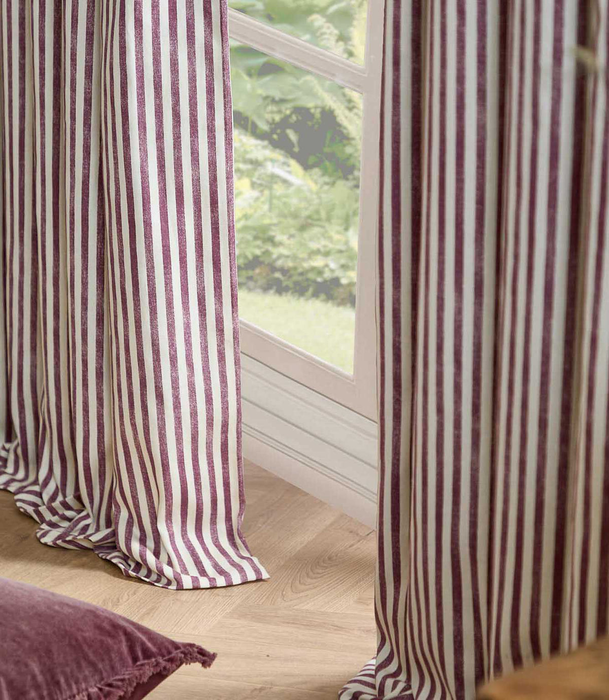 Striped curtains in a dusty burgundy and cream color, hanging on either side of a window in a room with light wood flooring.