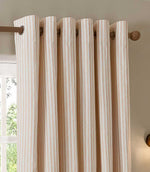 Close-up of blush pink and cream striped eyelet curtains hanging from a wooden curtain pole, showing the metal eyelets, neat folds and vertical stripe pattern.