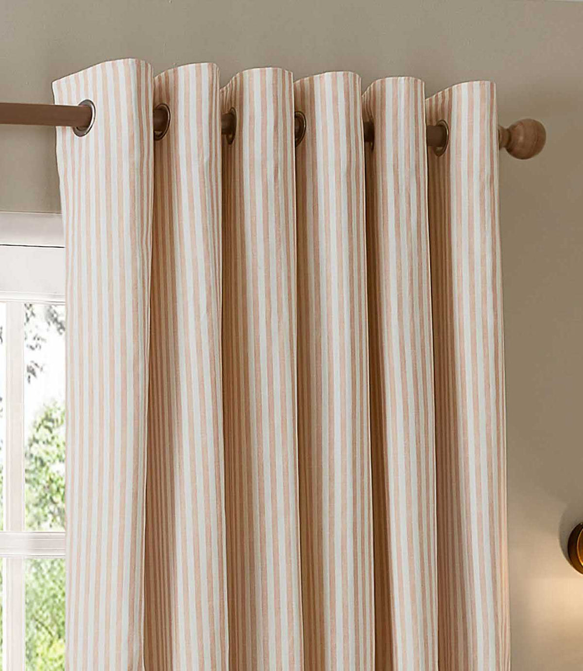 Close-up of blush pink and cream striped eyelet curtains hanging from a wooden curtain pole, showing the metal eyelets, neat folds and vertical stripe pattern.