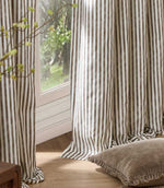 Brown and cream striped curtains hanging in soft full-length folds beside a bright window, styled with a neutral floor cushion and natural wood flooring.