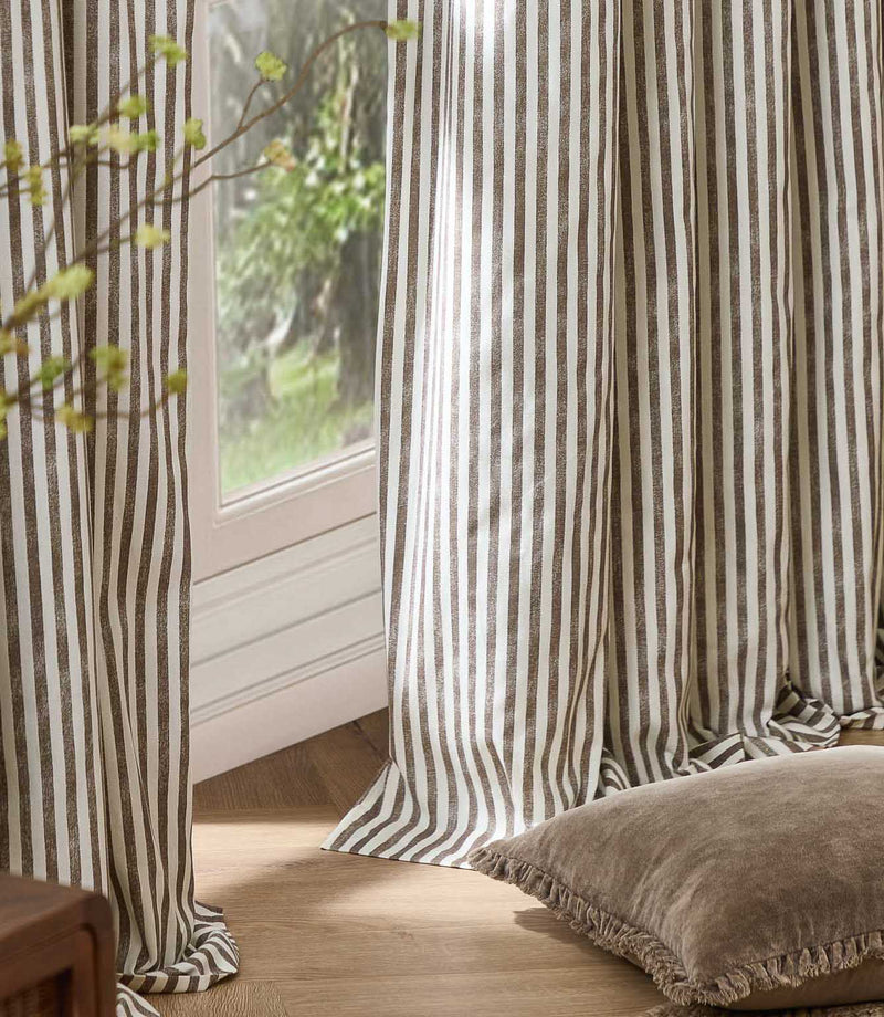 Brown and cream striped curtains hanging in soft full-length folds beside a bright window, styled with a neutral floor cushion and natural wood flooring.