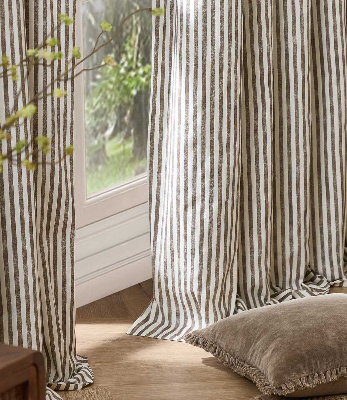 Brown and cream striped curtains hanging in soft full-length folds beside a bright window, styled with a neutral floor cushion and natural wood flooring.