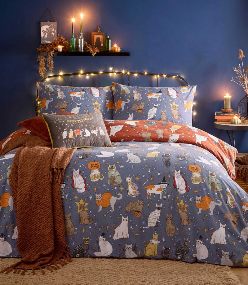 Halloween Pawty multicolour reversible duvet cover set with illustrated cats in witch, mummy, pumpkin and vampire costumes on a dusky blue background, styled with rust throws and cushions.