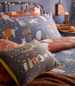 Close-up of Halloween Pawty reversible bedding showing costumed cats on dusky blue and rust backgrounds, styled with a grey Happy Halloween cat cushion and rust textured throw.
